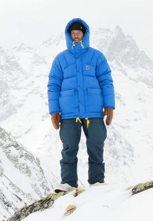 Blue puffer jacket with a hood, featuring yellow accents and two front pockets, paired with dark blue pants and brown gloves in a snowy landscape.
