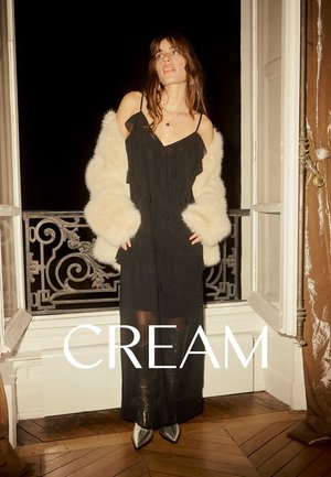 Schwarzes, gerafftes Kleid mit Spaghettiträgern, unter einem cremefarbenen Kunstfellmantel geschichtet. Durchsichtiger unterer Saum und spitze schwarze Stiefel.