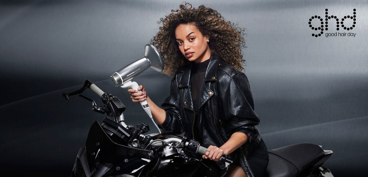 Femme aux cheveux bouclés portant une veste en cuir noire, tenant un sèche-cheveux, assise sur une moto noire devant un fond gris.