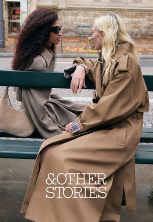 To kvinder iført brune trenchcoats sidder på en grøn bænk udendørs, vendt mod hinanden og i samtale.