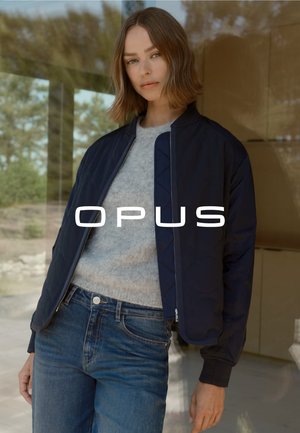Veste de pilote navy avec une texture matelassée, fermeture éclair argentée et poignets côtelés, portée sur un pull gris et associée à un jean bleu.