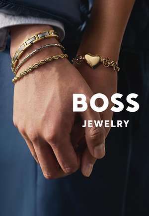 Zwei Hände mit goldenen Armbändern, eine mit einem herzförmigen Anhänger, auf dem "BOSS" eingraviert ist, vor einem dunklen Hintergrund mit dem Schriftzug "BOSS SCHMUCK".