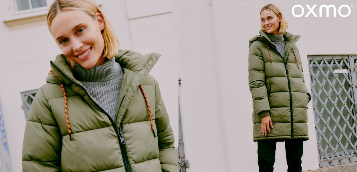 Doudoune vert olive avec fermeture éclair, capuche, et cordons de serrage orange contrastants. Portée sur un pull gris, associée à un pantalon noir.