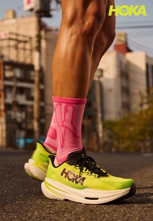 HOKA Laufschuhe in leuchtendem Grün mit schwarzen Akzenten, ausgestattet mit einer dicken Sohle. Der Träger trägt rosa gemusterte Socken, die über den Knöcheln sichtbar sind.