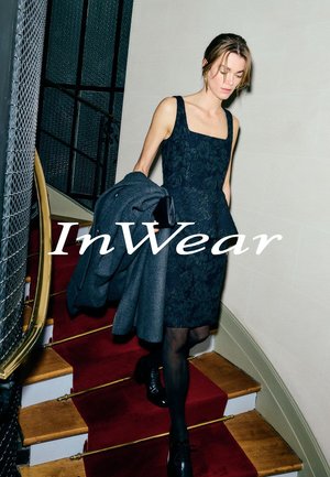 Abito nero in pizzo floreale con scollo quadrato, abbinato a un cappotto grigio, mentre scende una scala con moquette rossa.