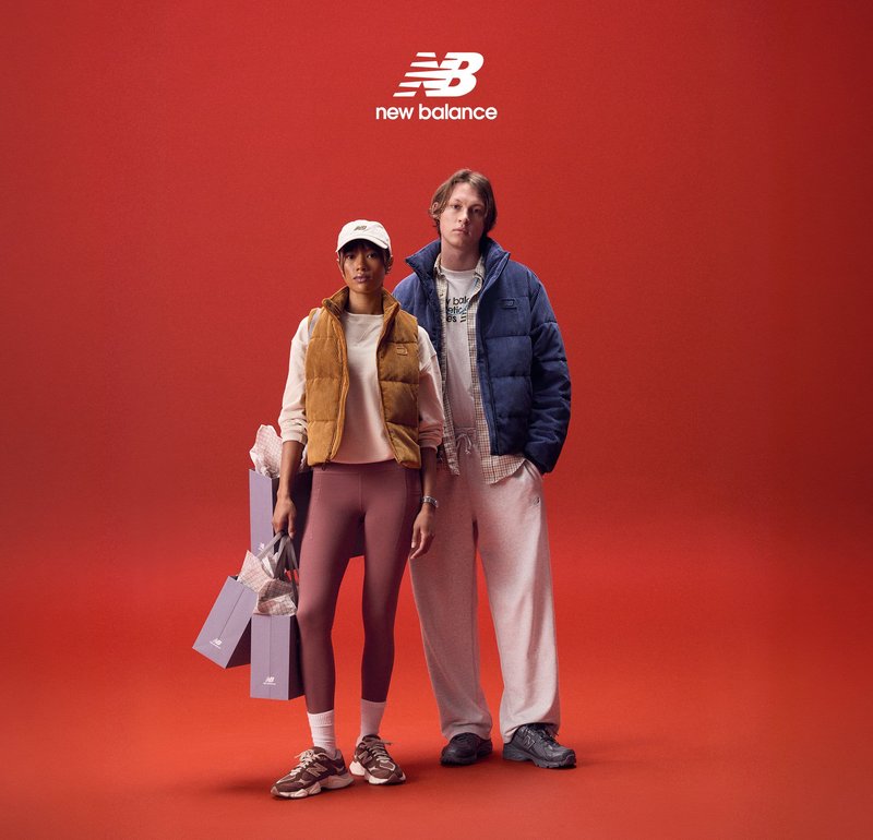 Due modelli sono in piedi davanti a uno sfondo rosso. Uno indossa un gilet imbottito giallo, una maglietta bianca, leggings marroni e sneakers; l'altro indossa una giacca imbottita blu e pantaloni della tuta grigi. Entrambi tengono borse della spesa.