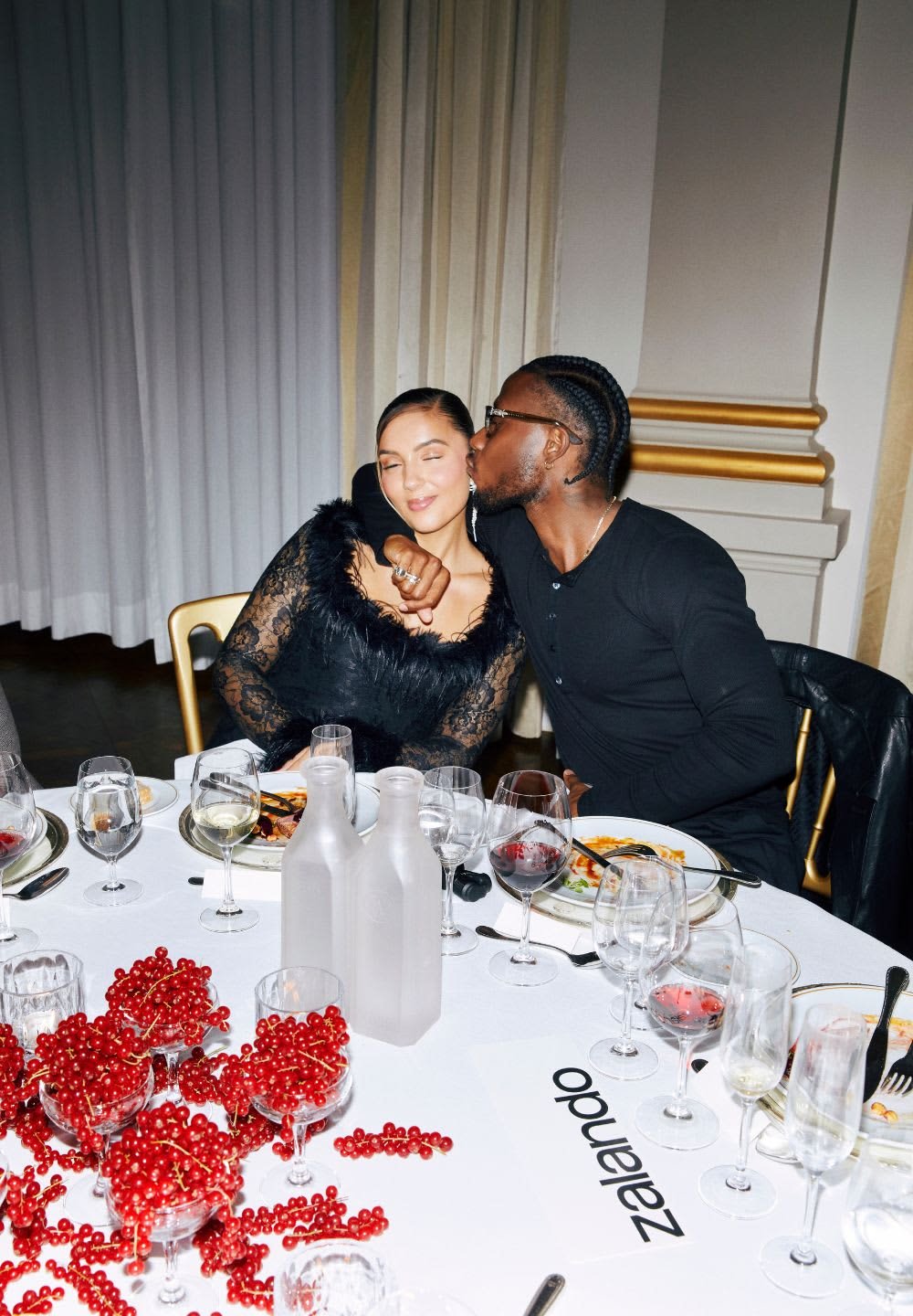 To personer ved et spisebord, den ene iført en sort top med pelsdetaljer, den anden i en sort langærmet skjorte. Bordet er dækket med tallerkener og glas.