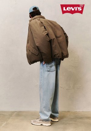 Brown padded jacket with a high collar, worn over light blue baggy jeans and beige sneakers. The person wears a light blue cap.