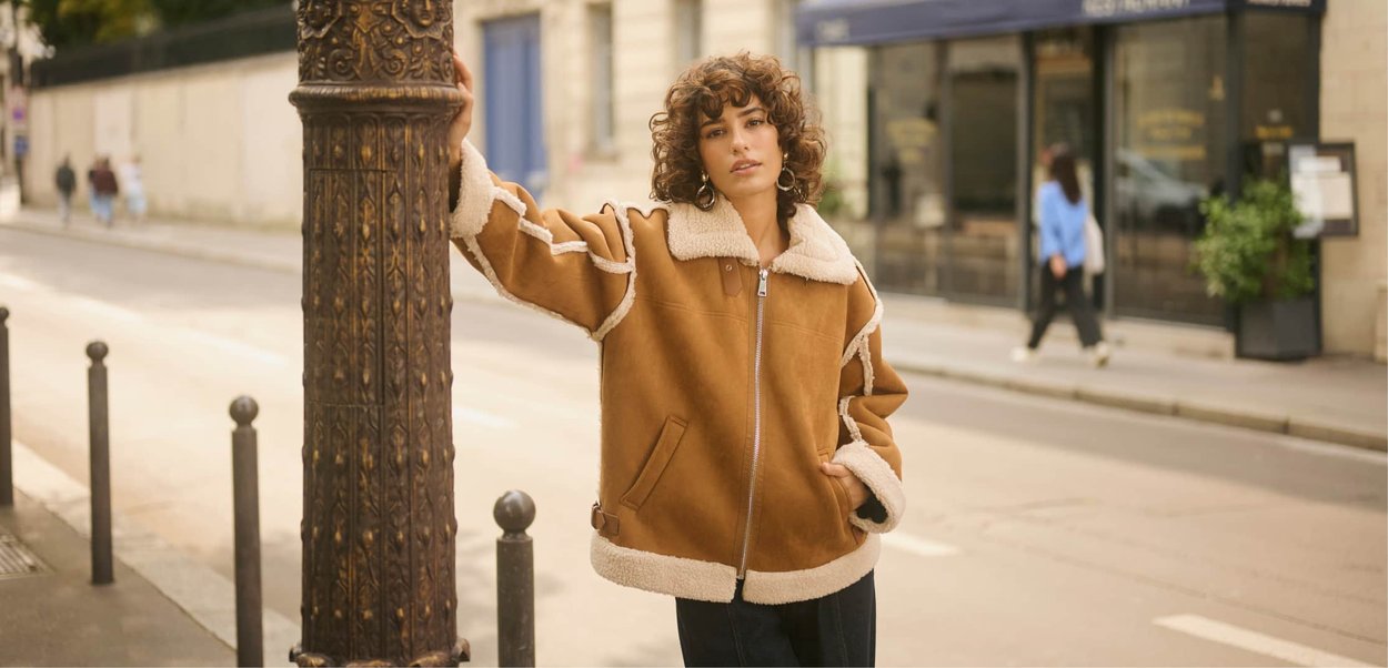 Tan faux shearling jacket with a white collar, large pockets, and a silver zipper, worn with black pants against an urban background.