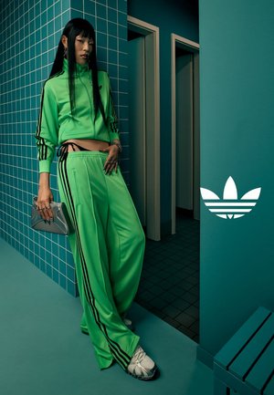 Groene trainingsoutfit gemaakt van glad materiaal, met zwarte strepen, een cropped top en losvallende broek. Inclusief een zilveren tas en witte sneakers.