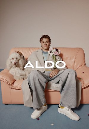 Das Model sitzt auf einer rosa Ledercouch, trägt einen hellgrauen Mantel, einen blauen Pullover und lockere Jeans. Es hält Blumen neben einem weißen Hund.