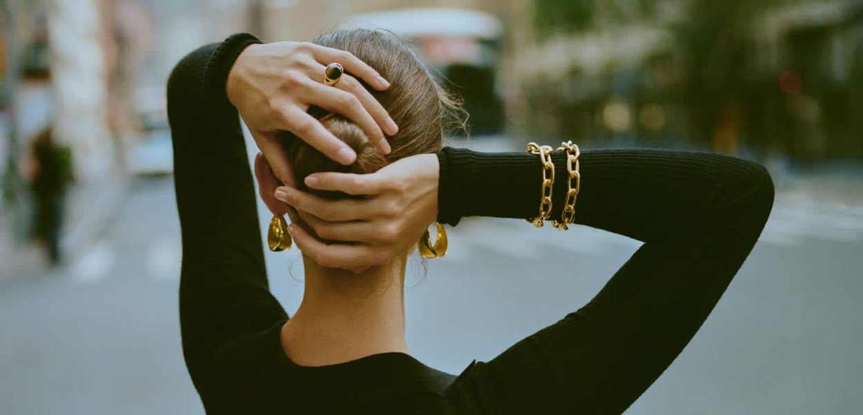 Des boucles d'oreilles en or, un bracelet en chaîne épaisse en or et une bague ornent les mains d'une personne, habillée d'un haut noir à manches longues ajusté. Les cheveux sont attachés.