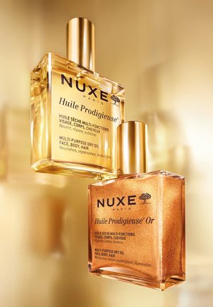 Deux flacons de NUXE Huile Prodigieuse, l'un transparent avec une huile jaune et des accents dorés, l'autre ambré avec des reflets scintillants, tous deux avec des bouchons dorés.