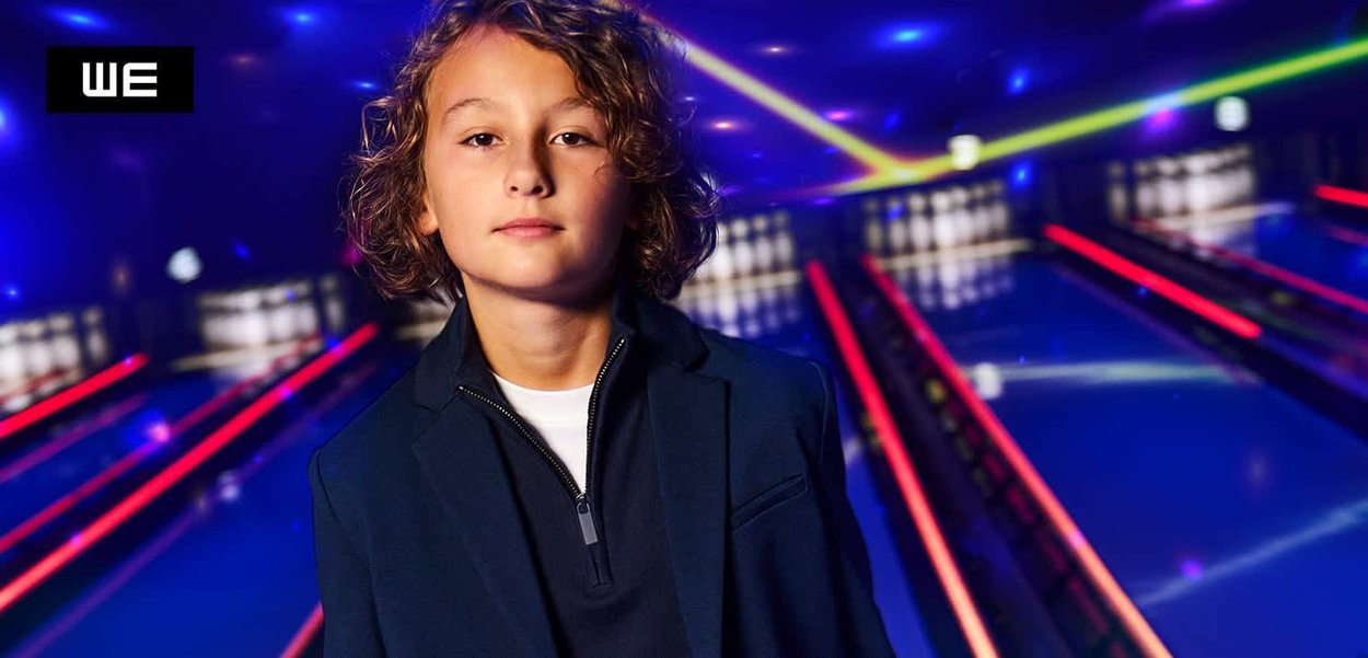 Zwart zip-up jack over een wit shirt, met lichtbruin krullend haar, tegen een kleurrijke bowlingbaan achtergrond met neonlichten.