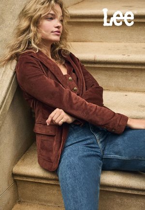 Bruine corduroy blazer met revers en knopen, gecombineerd met een blauwe spijkerbroek. Model zittend op trappen met zachte texturen en een neutrale achtergrond.