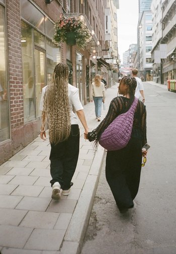 Twee personen lopen hand in hand op een straat van straatstenen. De een draagt een wit shirt en zwarte broek, de ander heeft een paarse gewatteerde tas en gevlochten haar.