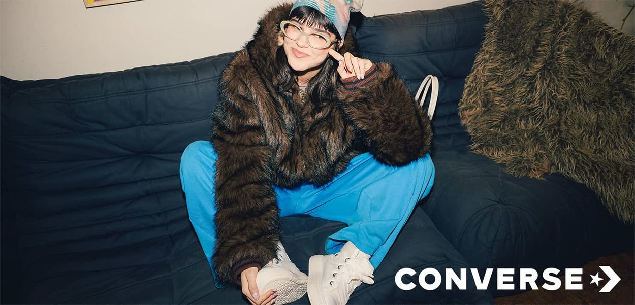 Giacca di pelliccia marrone, pantaloni della tuta blu e sneakers bianche alte. Seduto su un divano blu scuro con cuscini testurizzati.