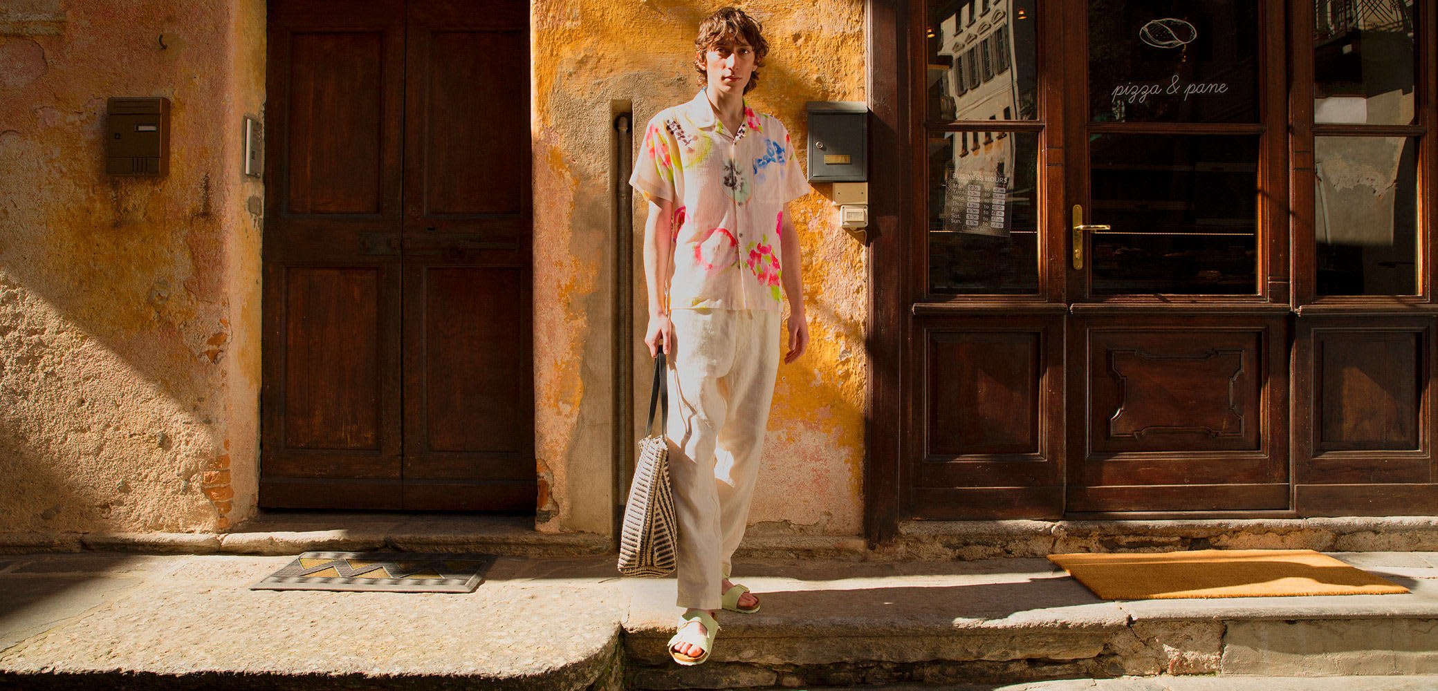 Camicia floreale a maniche corte nei toni pastello con accenti rosa e blu, pantaloni larghi color crema e borsa a tracolla con motivo.