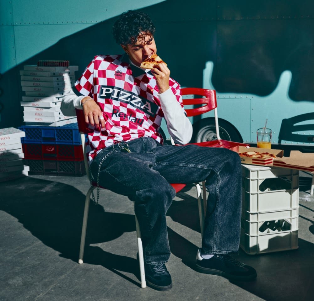 Camicia a maniche corte a quadretti rossi e bianchi con la scritta "PIZZA", jeans neri, seduto su una sedia, tiene in mano una fetta di pizza, con scatole di pizza dietro.
