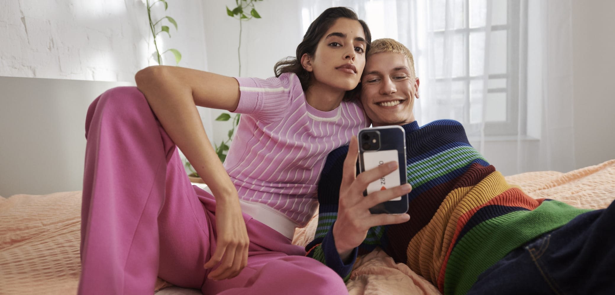 Due persone sono sedute su una coperta di un leggero rosa. Una indossa un maglione rosa a coste e pantaloni rosa, mentre l'altra indossa un maglione a righe multicolori.