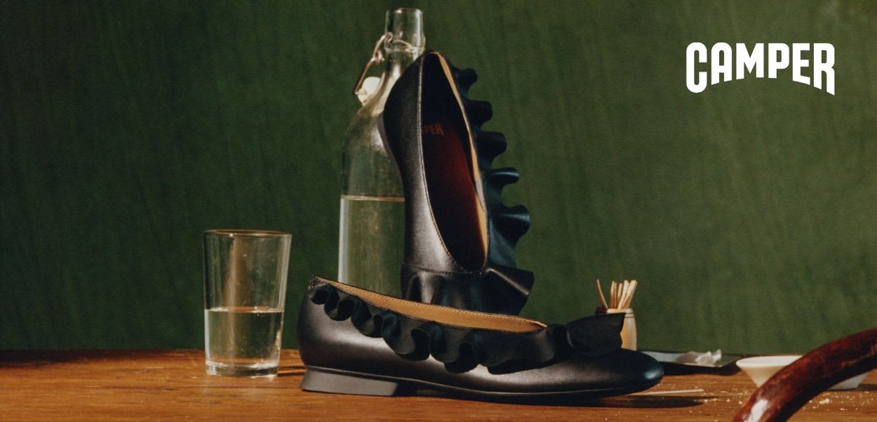 Zwarte leren damesschoenen met platte zool en gerimpelde rand, tentoongesteld op een houten tafel, met een glas, fles en luciferhouders op de achtergrond, groene achtergrond.