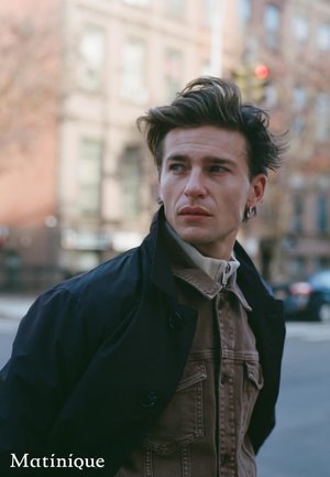 Junger Mann mit gestyltem dunklem Haar, trägt eine geschichtete Jacke und schaut auf einer Stadtstraße mit unscharfem Hintergrund von Gebäuden nach links.