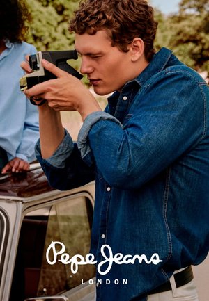 Jonge man in een denim jacket gebruikt een vintage camera buitenshuis nabij een klassieke auto, terwijl hij een foto maakt in een natuurlijke omgeving.