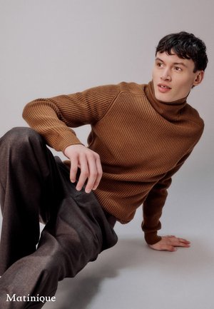 Brauner gerippter Rollkragenpullover mit eng anliegenden Ärmeln, kombiniert mit lockeren, dunkelgrauen Hosen. Das Model sitzt lässig vor einem hellen Hintergrund.