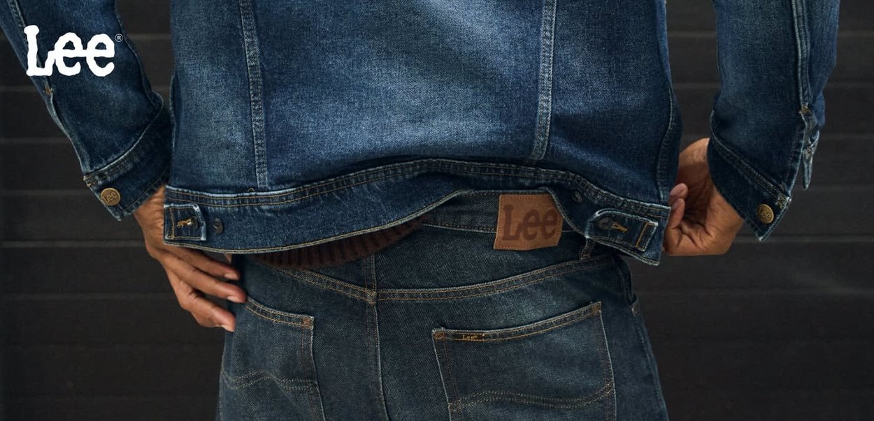 Tummansinisestä denim-takista, jossa on vahvistettu kaulus, nappikiinnitys hihansuissa ja ruskea nahkainen Lee-logo-läppä vastaavien farkkujen vyötäröllä.