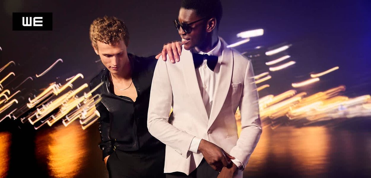 Due uomini posano insieme di notte. Uno indossa una camicia nera; l'altro indossa un frac bianco con papillon. Lo sfondo presenta luci sfocate.