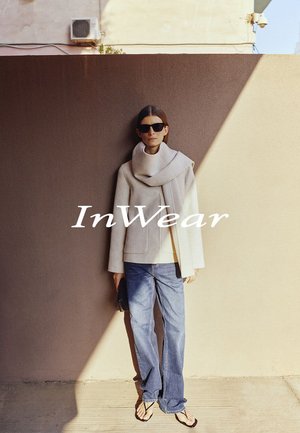 Frau trägt übergroßen beigen Schaljacke, blaue Jeans und schwarze Sonnenbrille, steht vor einer zweifarbigen Wand mit Sonnenlicht und Schatten.