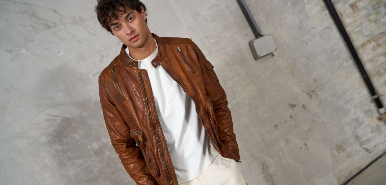 Giacca in pelle marrone con dettagli di cerniera e tasche, indossata sopra una camicia bianca. Texture morbida, design aderente e stile casual.