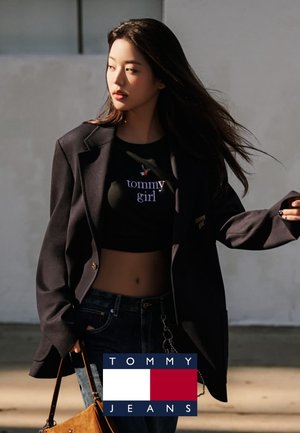 Top corto negro con texto "tommy girl", americana azul marino y jeans oscuros. Bolso marrón con detalle de cadena. Logo de Tommy Jeans en la esquina.
