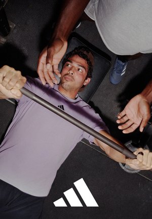 Barre avec poignées noires, tenue par une personne portant un t-shirt Adidas violet clair, tandis qu'une autre main apporte de l'assistance d'en haut. Sol de gymnase noir visible.