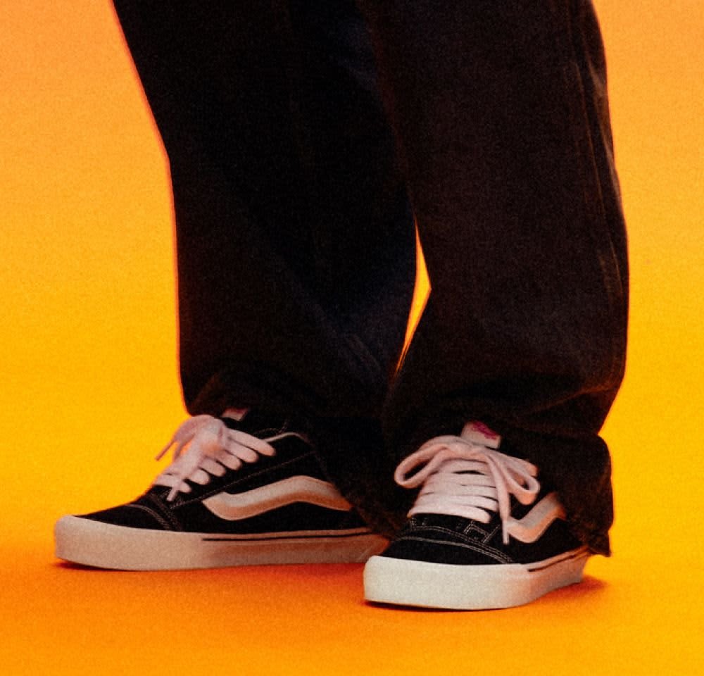 Schwarze Canvas Low-Top Sneaker mit weißen Schnürsenkeln, einem weißen Streifenmuster und einer weißen Gummisohle. Getragen mit dunklen Jeans auf einem orangefarbenen Hintergrund.