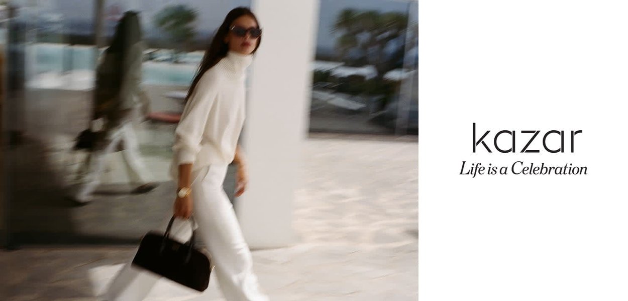 Model wearing a cream turtleneck sweater and white trousers, carrying a black handbag. Background features a blurred outdoor area with greenery.
