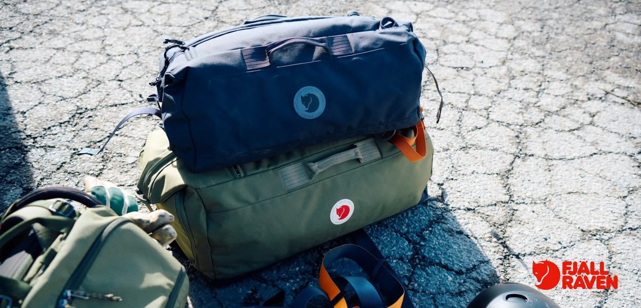 Zwei Fjällräven Reisetaschen übereinander gestapelt, eine in Dunkelblau mit Griffen, die andere in Olivgrün mit runder Logo und orangefarbenem Akzent. Strukturierten Oberfläche.