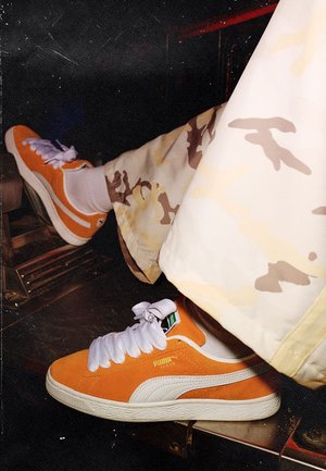 Persoon draagt oranje en witte Puma Suede sneakers met witte veters en beige camouflagebroeken, rust de voeten op een metalen oppervlak.
