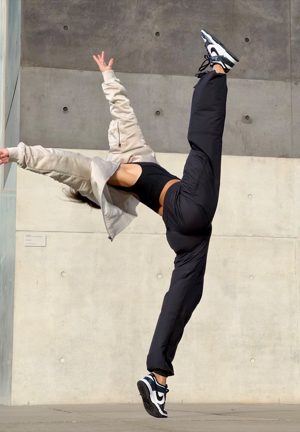 Black athletic pants, cropped black tank top, and beige oversized jacket. Black and white sneakers. Leg extended in a dynamic pose.