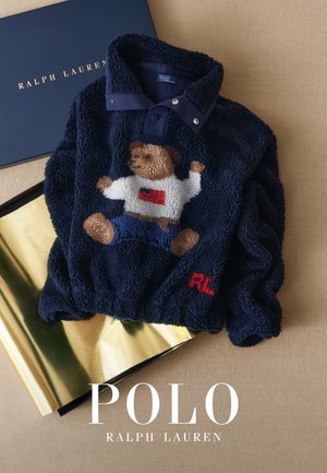 Marineblå fleecegenser med et bjørnemotiv på forsiden, med krage og rødt brodert logo. Gull- og svart bakgrunn.