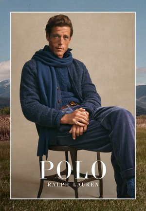 Man zit op een stoel en draagt gelaagde blauwe breiwerk, een sjaal en corduroy broek, met de tekst "Polo Ralph Lauren" onderaan.