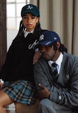 Deux personnes portant des casquettes de baseball et des vêtements décontractés preppy, l'une posant la main sur l'épaule de l'autre à l'intérieur sous un éclairage doux.