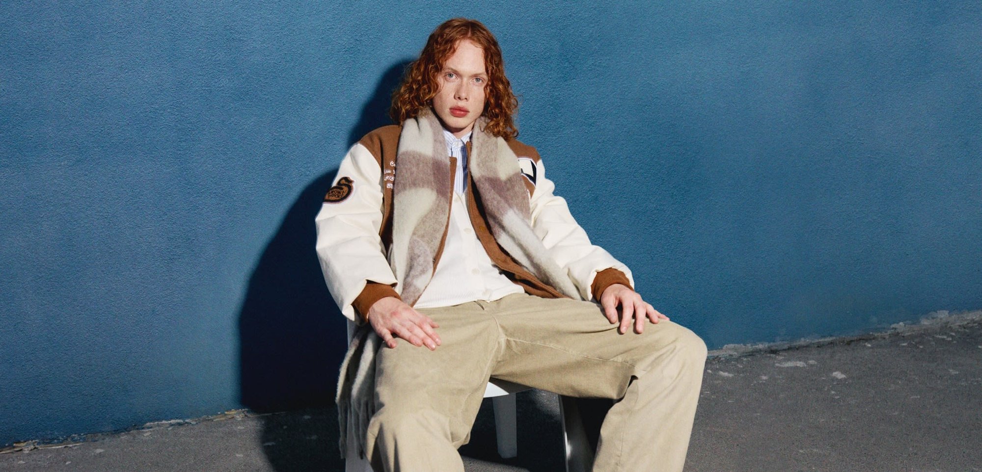 Corduroy pants, white shirt, brown bomber jacket with embroidered details, and patterned scarf. Dark shoes, seated on a white chair against a blue wall.