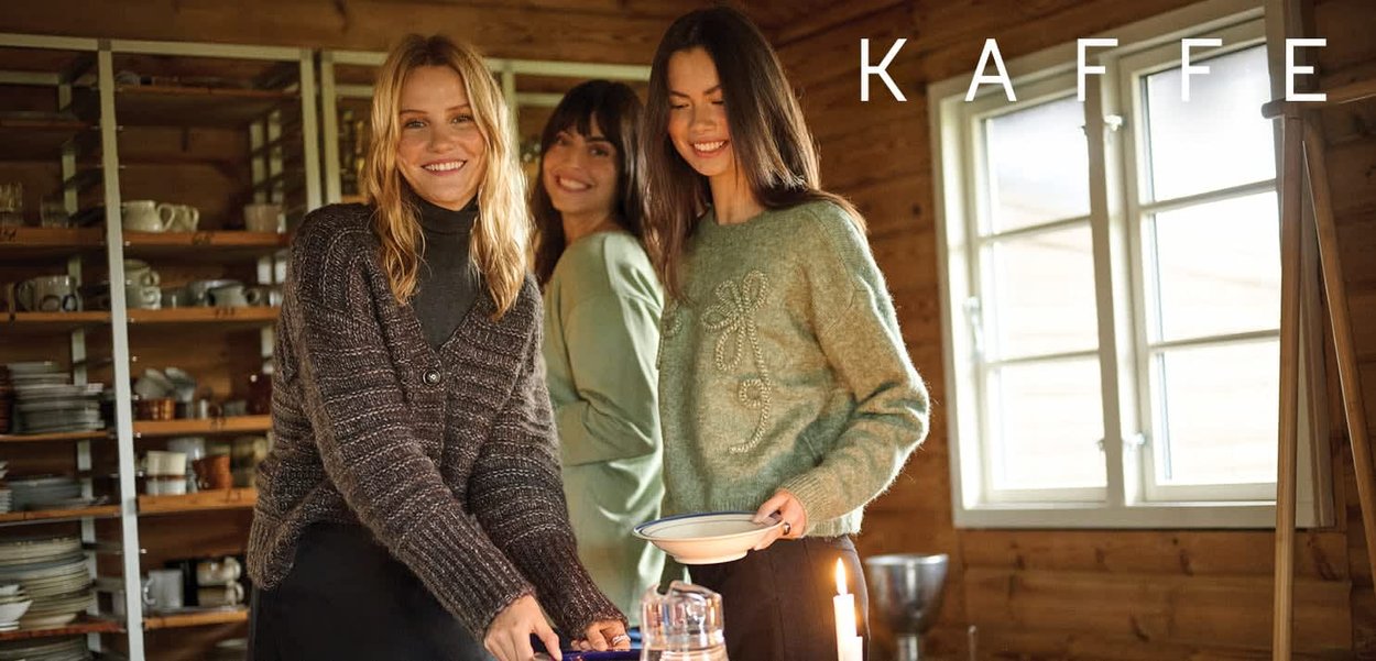 Tre kvinder i sweatere står i et trærum, en holder en skål, der fremviser teksturerede strik i mørke og lyse farver. Hylder viser service.