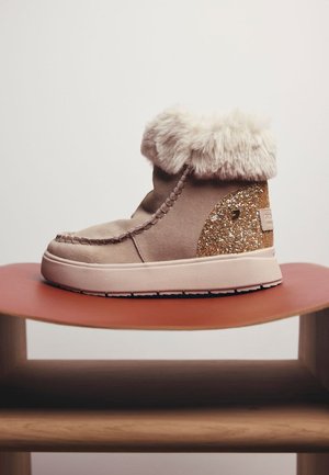 Stivale alla caviglia beige con risvolto soffice, materiale scamosciato, cuciture decorative e una pannello di accentuazione dorato glitterato sul tallone. Suola piatta in gomma.