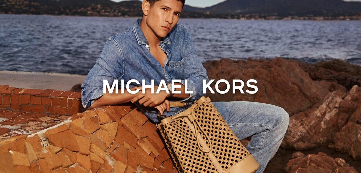 Mann in Jeanshemd und Jeans sitzt auf Terrakottaplatten am Meer und hält eine hellbraune, perforierte Lederhandtasche von Michael Kors.