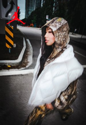 Un jeune portant un sweat à capuche et un pantalon camouflage, avec un gilet rembourré blanc, se promène dans une rue urbaine, proche de panneaux de circulation et de bâtiments.