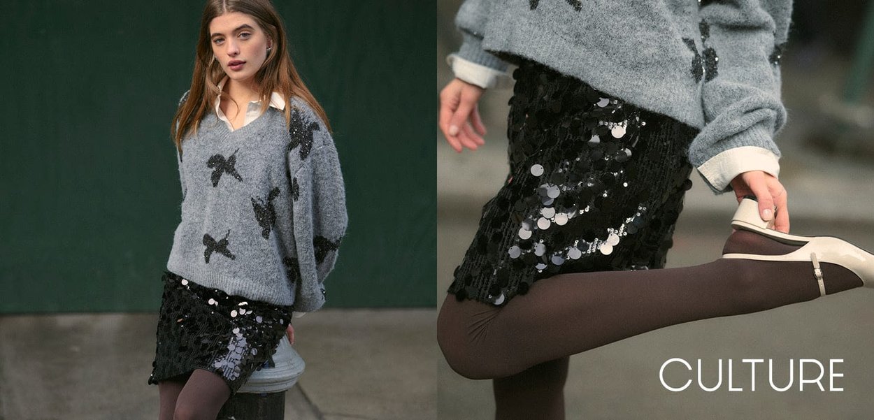Grigia maglietta con design a fiocco neri, abbinata a una gonna nera con paillettes. Il modello indossa collant marroni e décolleté color crema, evidenziando la consistenza e la lucentezza dell'outfit.