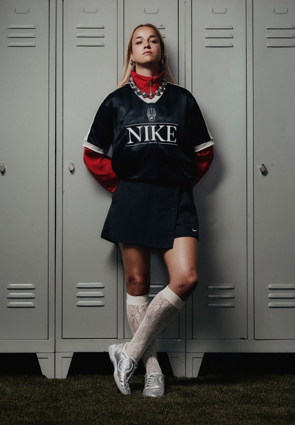Nike sports jersey in black with white accents, paired with a red long-sleeve shirt, dark skirt, lace socks, and silver sports shoes against lockers.