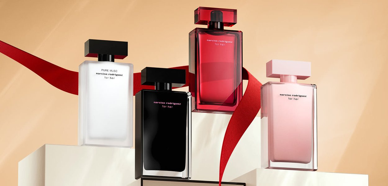 Quattro flaconi di profumo su un piedistallo bianco: vetro trasparente con accenti neri e rosa, con forme diverse e tappi distintivi. Sfondo con nastro rosso.